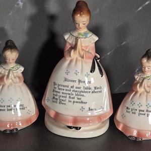 May include: Three ceramic figurines of children with hands clasped in prayer. Each wears a white dress with a pink hem and blue floral accents. The central figure features the text Dinner Prayer, while the side figures display prayer text.