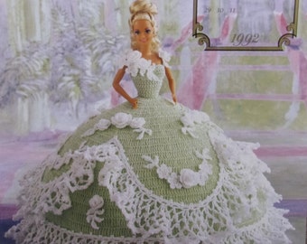 May include: A doll in a light green crocheted dress with white lace trim and floral details. The doll has blonde hair and is positioned in front of a 1992 calendar. The dress has a full skirt and a detailed bodice.