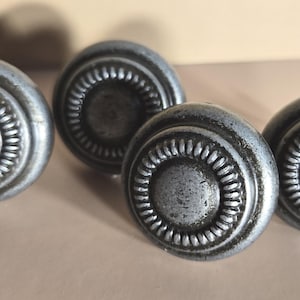 May include: Four round cabinet knobs with a dark, weathered silver finish. Each knob features a smooth, raised center disc surrounded by a textured, ridged border. The hardware has a matte, metallic appearance with subtle dark shading.