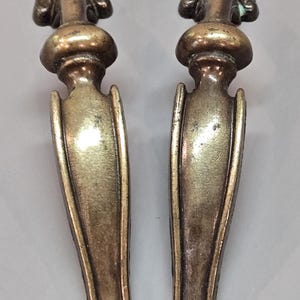 May include: A pair of matching antique brass drawer pulls featuring a vertical elongated design with a rounded top section, a central bulbous element, and a curved handle body that terminates in a small circular base at the bottom end.
