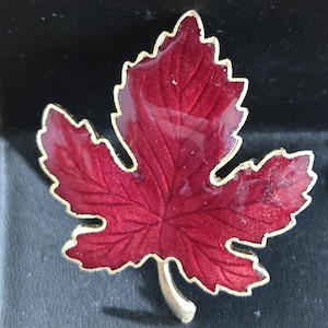 May include: A maple leaf brooch featuring a deep red enamel finish with a gold-toned metal border. The leaf design includes detailed embossed veins across the surface and a small stem at the bottom, creating a realistic botanical appearance.