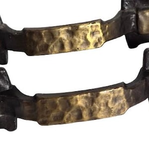May include: Two antique-style cabinet pulls. The pulls have a dark bronze finish with a textured, hammered brass-colored center section. Each pull has a rectangular end with a decorative design.