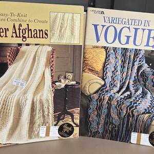 May include: Two softcover craft books titled Sampler Afghans and Variegated in Vogue. The first book features a cream-colored textured knit blanket, while the second displays a multi-colored crocheted afghan with fringe on a yellow sofa.