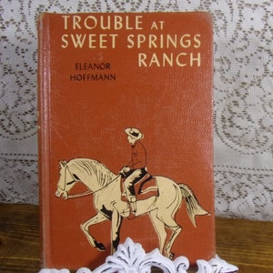 May include: A vintage book titled "Trouble at Sweet Springs Ranch" by Eleanor Hoffmann. The cover is a reddish-orange color with an illustration of a person riding a horse. The book is displayed on a white decorative stand.