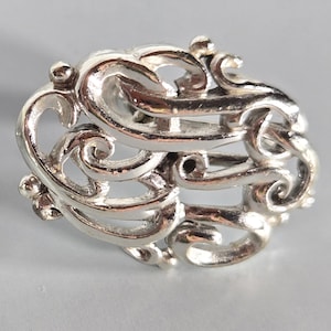 May include: A polished silver brooch featuring an intricate, openwork design of swirling, interconnected metal scrolls and loops. The metallic surface reflects light, highlighting the curved, organic shapes and small spherical accents.