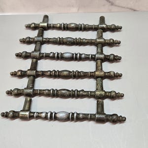 May include: Six metal drawer pulls arranged in a grid. Each pull features a central spindle shape with decorative rings and tapered ends, finished in a weathered, antiqued metallic tone with dark patina and highlights of silver and gold.