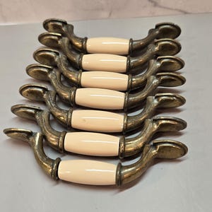 May include: A set of six drawer pulls arranged in a vertical stack. Each pull features a central, smooth, off-white ceramic handle section flanked by curved, aged brass-toned metal ends with circular mounting plates at the outer edges.