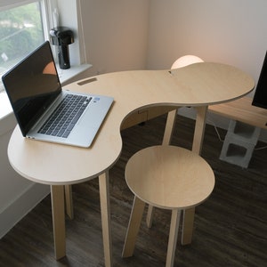 May include: A light wood desk with a curved top and a matching stool. The desk has a laptop on it and a lamp on the side. The desk and stool are on a dark wood floor.