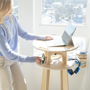 May include: A woman works at a standing desk with a laptop and phone. The desk is made of light wood and has a shelf for storage. The desk is in front of a window with a view of a city.