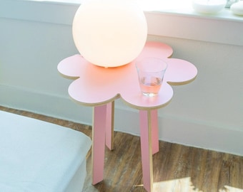 May include: A pink flower-shaped side table with four legs. A white globe lamp sits on the table, and a glass of liquid is on the table to the right of the lamp.