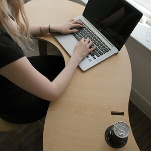 May include: A person sits at a light wood desk with a laptop computer and a can of soda. The desk is curved and has a unique design.