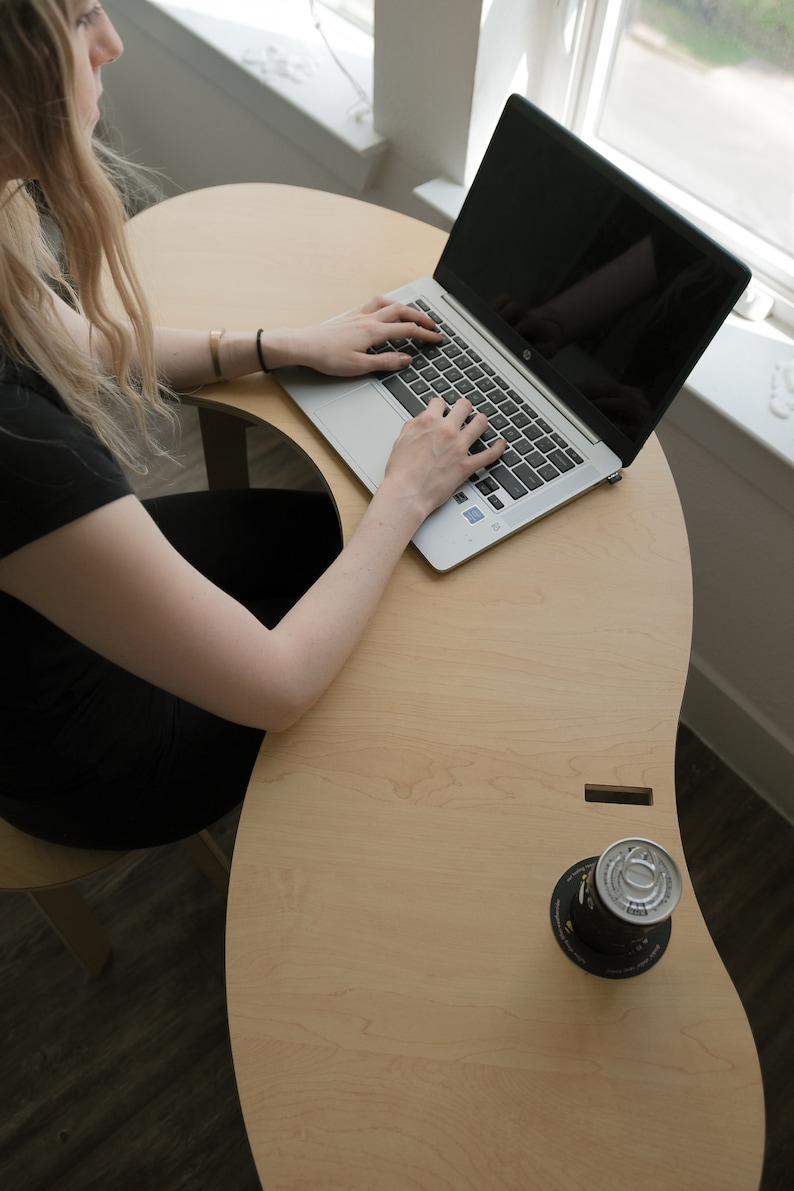 May include: A person sits at a light wood desk with a laptop computer and a can of soda. The desk is curved and has a unique design.