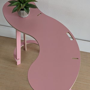 May include: A pink, curved table with a unique design. The table has a small shelf on the side and a cutout on the top surface. A small potted plant sits on the table.