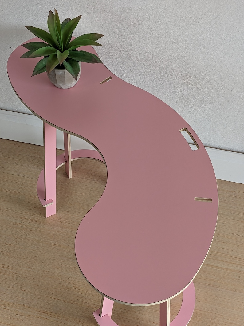 May include: A pink, curved table with a unique design. The table has a small shelf on the side and a cutout on the top surface. A small potted plant sits on the table.