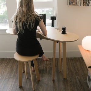 May include: A woman sits at a light wood desk with a laptop and a can of soda. The desk has a unique curved shape and is supported by four legs. The desk is paired with a matching stool.