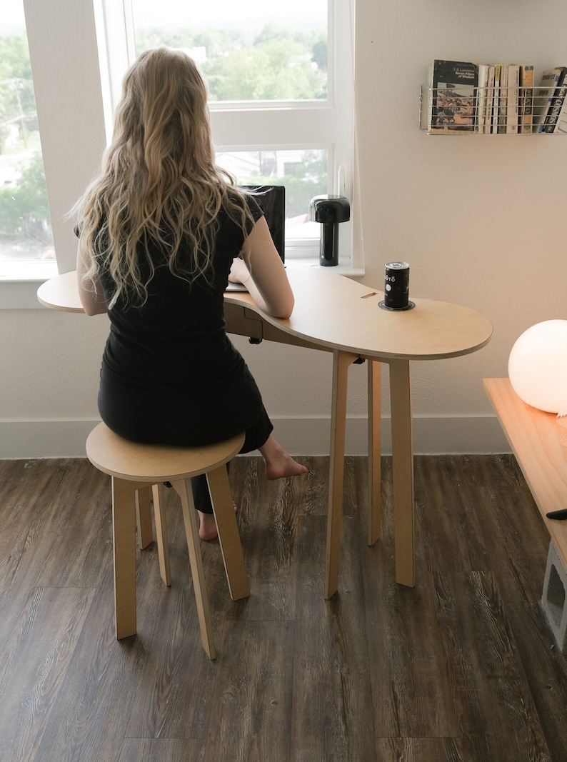 May include: A woman sits at a light wood desk with a laptop and a can of soda. The desk has a unique curved shape and is supported by four legs. The desk is paired with a matching stool.
