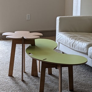 May include: Two wooden tables with unique shapes. The larger table is green and shaped like a kidney bean. The smaller table is brown and shaped like a flower. Both tables have four legs.