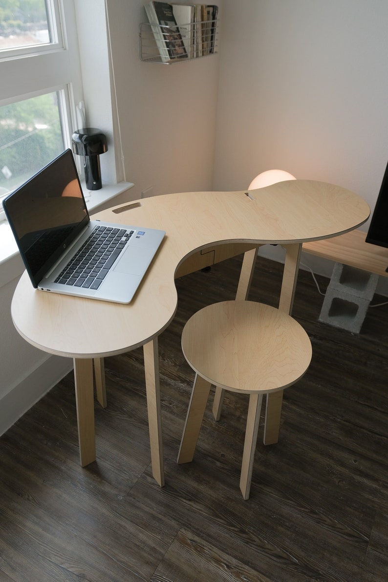 May include: A light wood desk with a curved top and a matching stool. The desk has a laptop on it and a lamp on the side. The desk and stool are on a dark wood floor.