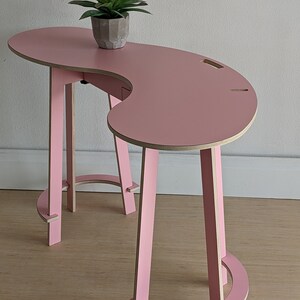 May include: A pink, curved desk with a unique, modern design. The desk has a single leg on one side and two legs on the other side. The legs are made of wood and are painted pink. The desk is made of plywood and has a smooth, pink finish. A small potted plant sits on the desk.