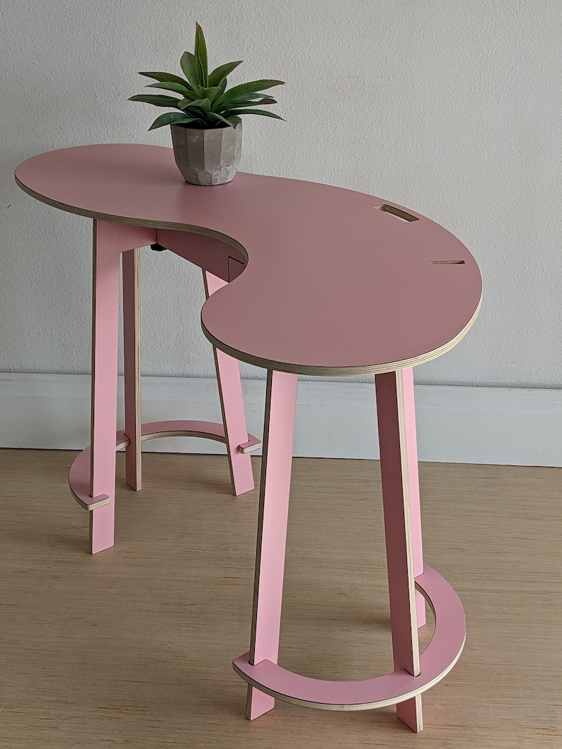May include: A pink, curved desk with a unique, modern design. The desk has a single leg on one side and two legs on the other side. The legs are made of wood and are painted pink. The desk is made of plywood and has a smooth, pink finish. A small potted plant sits on the desk.