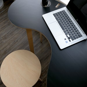 May include: A black, curved desk with a laptop computer and a can of soda on a coaster. A small, round, light wood stool is in the foreground.