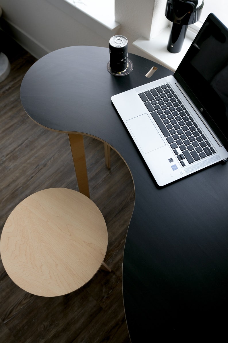 May include: A black, curved desk with a laptop computer and a can of soda on a coaster. A small, round, light wood stool is in the foreground.