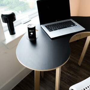 May include: A black laptop computer sits on a black, oval-shaped table with wooden legs. A can of "SOTO" sake sits on a coaster on the table.