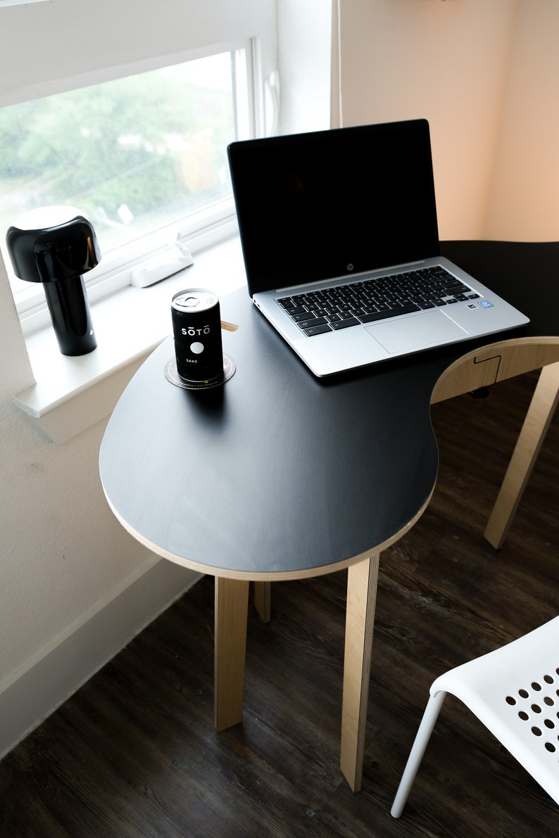 May include: A black laptop computer sits on a black, oval-shaped table with wooden legs. A can of "SOTO" sake sits on a coaster on the table.