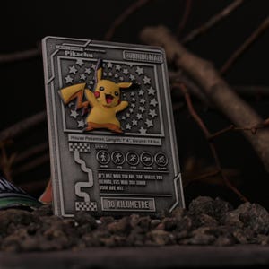 May include: A silver metal card featuring a Pikachu Pokemon character graphic. The card has the text "RUNNING MAD" and "10 KILOMETRE" on it. The card is resting on a bed of small grey stones.