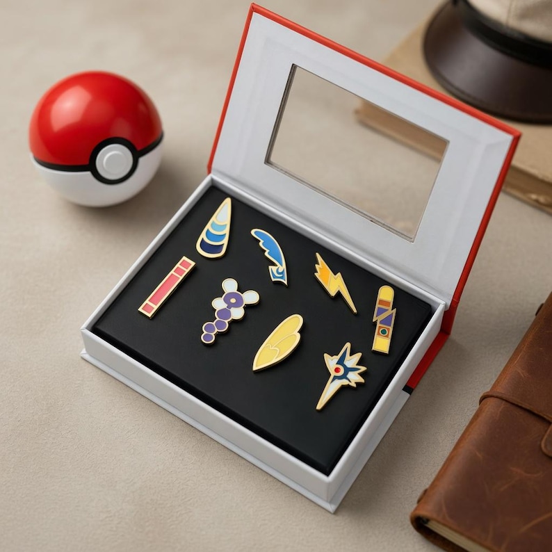 May include: A set of eight colourful enamel pins arranged inside a white display box with a clear window lid. The pins feature various abstract shapes including a lightning bolt, a wing, and geometric patterns. A red and white Pok&eacute; Ball sits nearby.