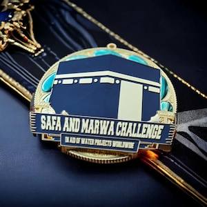 Può includere: Medaglia dorata con un designo blu e oro. La medaglia presenta una silhouette nera della Kaaba, un edificio a forma di cubo a La Mecca, Arabia Saudita. Il testo "Safa and Marwa Challenge" è scritto in lettere dorate su uno stendardo nero. Il testo "In aid of water projects worldwide" è scritto in lettere dorate più piccole sotto lo stendardo.