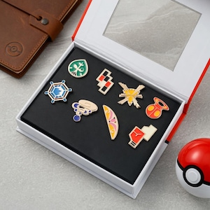 May include: A collection of eight colourful metal enamel pins arranged inside a white display box with a clear window. The pins feature various geometric and abstract shapes in shades of green, blue, yellow, red, pink, and black. A brown leather notebook and a red and white spherical toy are positioned nearby.