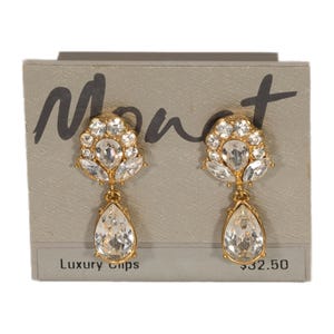 May include: A pair of gold-toned clip-on earrings with clear, sparkling crystals. The earrings feature a floral design at the top and a teardrop-shaped crystal pendant. The earrings are displayed on a gray card with the brand name "Monet".