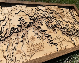 Op de afbeelding: Een houten kaart van het Grand Canyon National Park, met een kompasroos in het midden en de woorden "Grand Canyon National Park" eronder gedrukt. De kaart is gegraveerd met ingewikkelde details van de topografie van de canyon, waaronder het Kaibab-plateau, het Hualapai Indian Reservation en het Coconino-plateau.