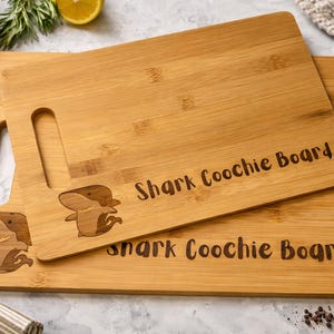 May include: Two rectangular bamboo cutting boards. The top board has a handle and the words "Shark Coochie Board" engraved. A cartoon shark is also engraved on the board. The bottom board has the same text and shark design.