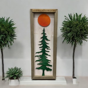 Pode incluir: Arte de parede com moldura de madeira com uma silhueta de árvore verde, um sol laranja e o texto "not all those who wander are lost". A obra de arte tem um tema rústico e ao ar livre.