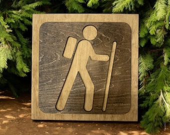 Pode incluir: Placa de caminhada de madeira com um recorte de uma pessoa com mochila e bengala. A placa tem um fundo marrom escuro com uma borda e uma figura marrom mais clara. A placa é exibida em um cenário de folhagem verde.