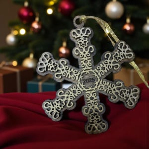May include: A wooden snowflake Christmas ornament with intricate cut-out designs and the year "2025" in the center. The ornament is brown with gold accents and hangs from a gold ribbon. It rests on a red surface.