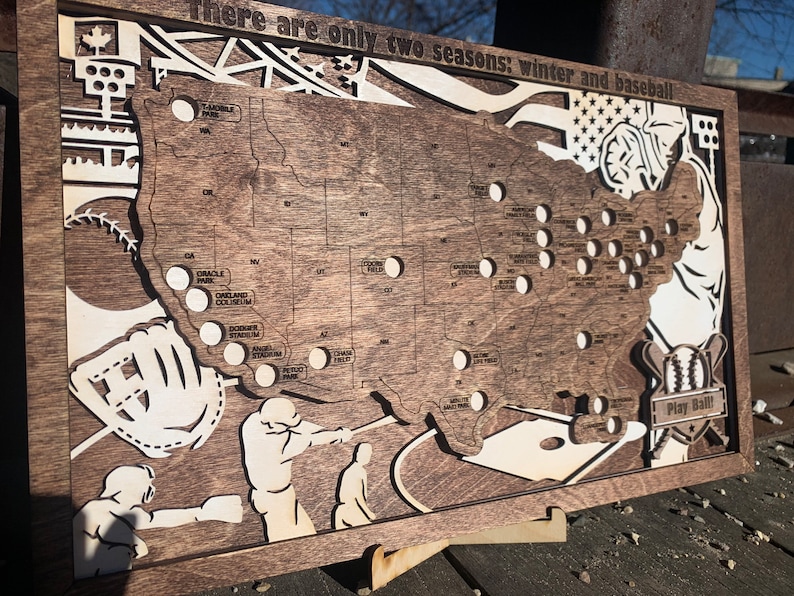 May include: A wooden map of the United States with baseball stadiums marked. The map is brown with white outlines of the states and stadiums. The text "There are only two seasons: winter and baseball" is at the top of the map. The text "Play Ball!" is at the bottom of the map.