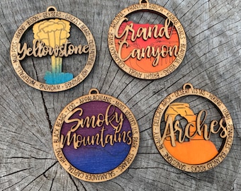 May include: Four wooden ornaments with a nature theme. Each ornament features a different national park: Yellowstone, Grand Canyon, Smoky Mountains, and Arches. The ornaments are painted with a gradient of colors, and each has a circle of text around the edge listing features of the park.