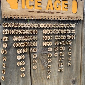 May include: A wooden sign with the words "The Ice Age National Scenic Trail" and an outline of the state of Wisconsin. The sign has a series of wooden circles hanging from it, each with a different name of a location on the trail.