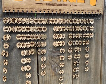 May include: A wooden sign with the words "The Ice Age National Scenic Trail" and an outline of the state of Wisconsin. The sign has a series of wooden circles hanging from it, each with a different name of a location on the trail.