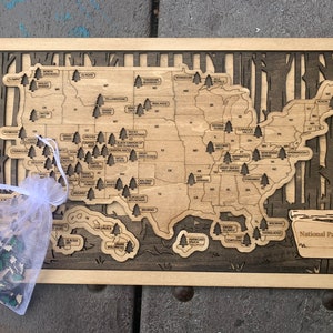 May include: A wooden map of the United States with black wooden trees marking the location of each national park. The map is framed in a light wood frame. A small white bag with green trees is in the lower left corner of the image. The text "National Parks" is in the lower right corner of the map.