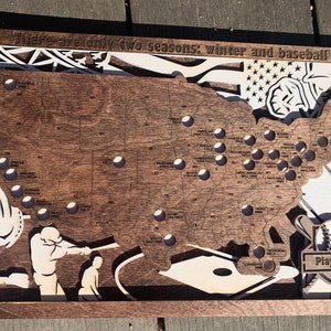 May include: A wooden map of the United States with a baseball theme. The map is decorated with baseball bats, a glove, and a baseball. The text "There are only two seasons: winter and baseball" is at the top of the map. The text "Play Ball!" is at the bottom of the map.