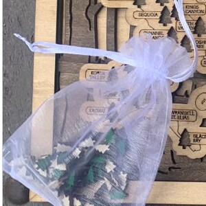 May include: A white mesh bag filled with small wooden puzzle pieces shaped like pine trees. The pieces are green, brown, and white. The bag is sitting on a wooden surface with a map of the United States.