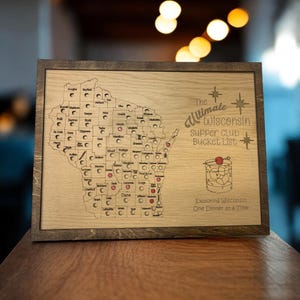 May include: A wooden map of Wisconsin with red dots marking locations. The text "The Ultimate Wisconsin Supper Club Bucket List" is printed on the map. The map is framed in a dark wood frame.