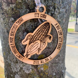 Puede incluir: Adorno de madera con dise&ntilde;o de mazorca de ma&iacute;z y cinta plateada. El adorno presenta una ilustraci&oacute;n de mazorca de ma&iacute;z y el texto "It's Corn", junto con las frases "A big lump with knobs", "It has the juice" y "I can't imagine a more beautiful thing".