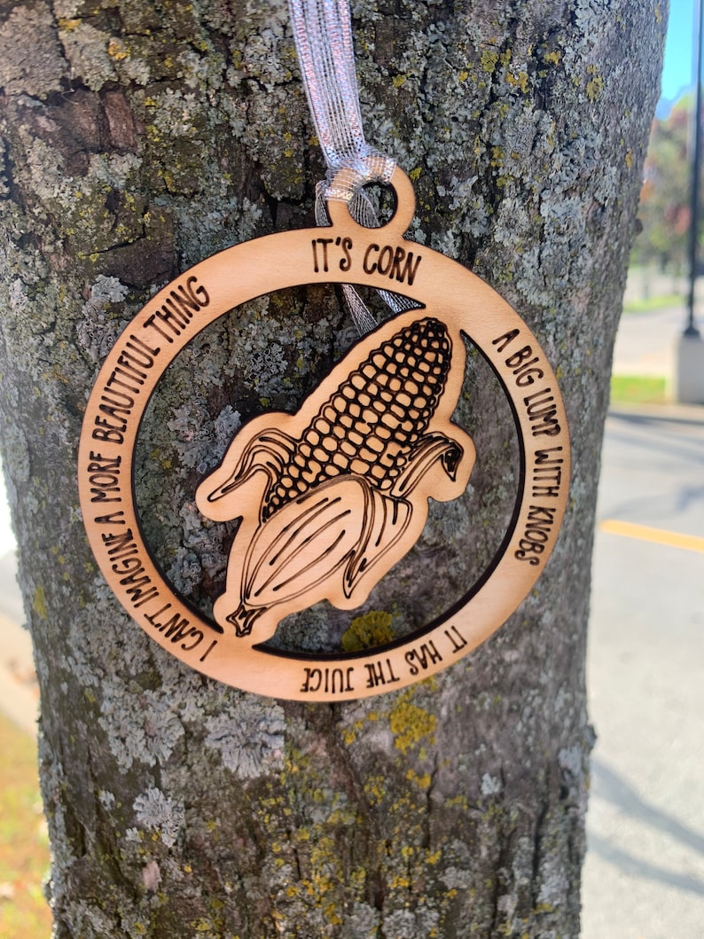 Puede incluir: Adorno de madera con dise&ntilde;o de mazorca de ma&iacute;z y cinta plateada. El adorno presenta una ilustraci&oacute;n de mazorca de ma&iacute;z y el texto "It's Corn", junto con las frases "A big lump with knobs", "It has the juice" y "I can't imagine a more beautiful thing".