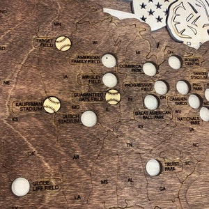 May include: A wooden map of the United States with baseball stadiums marked with baseballs. The map is stained a dark brown color and has a distressed look. The stadiums are marked with white circles and the names of the stadiums are printed in black.