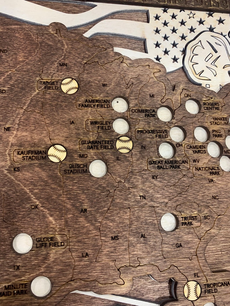 May include: A wooden map of the United States with baseball stadiums marked with baseballs. The map is stained a dark brown color and has a distressed look. The stadiums are marked with white circles and the names of the stadiums are printed in black.